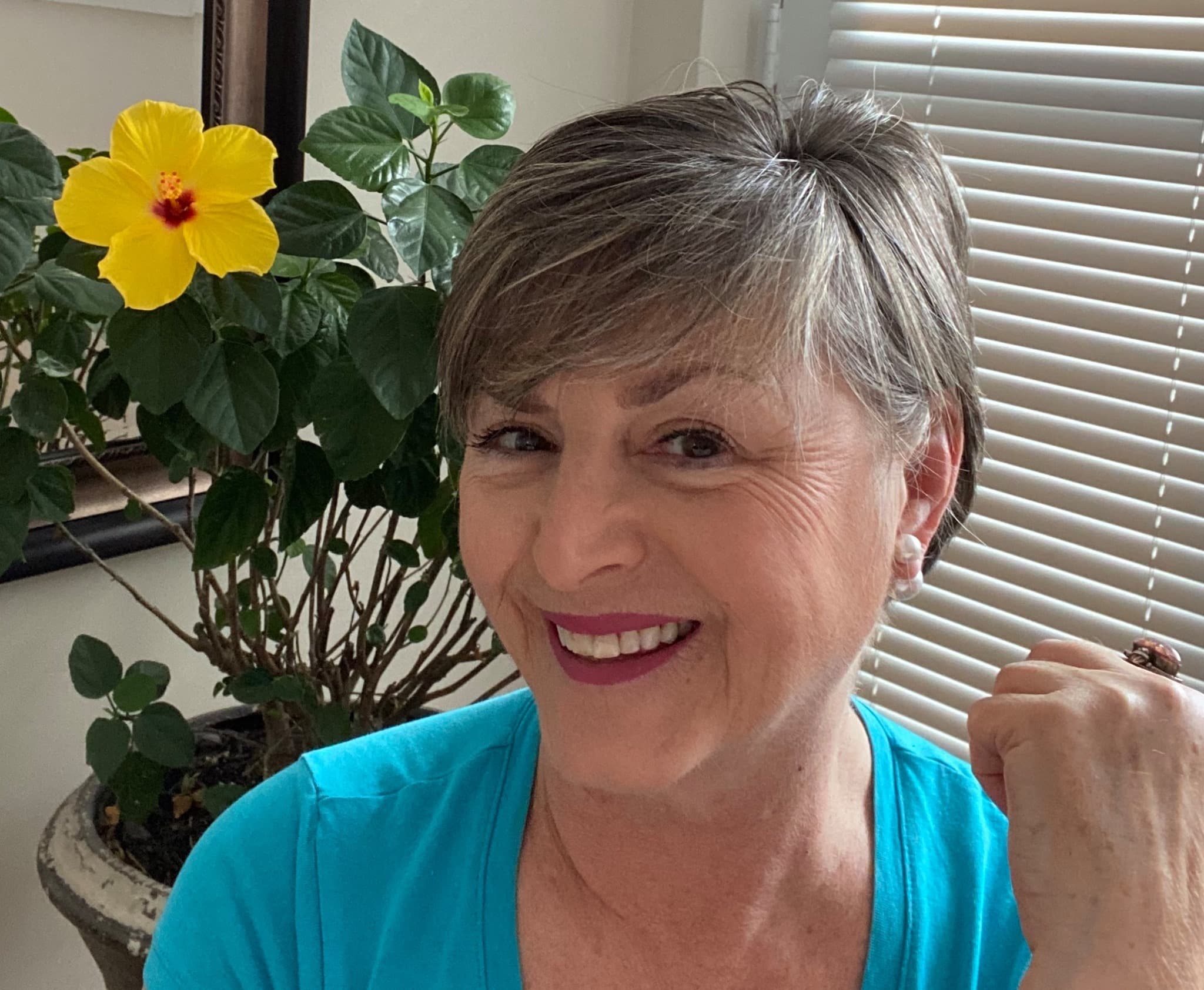 Smiling woman with short grey hair in a blue shirt beside a yellow hibiscus flower.