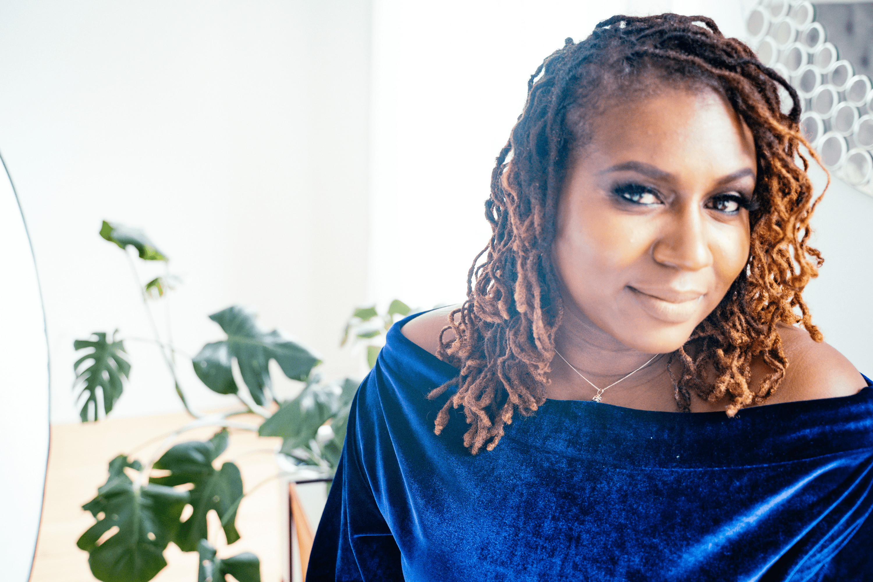Smiling Black woman with long brown dreadlocks wearing a blue velvet off-the-shoulder top.